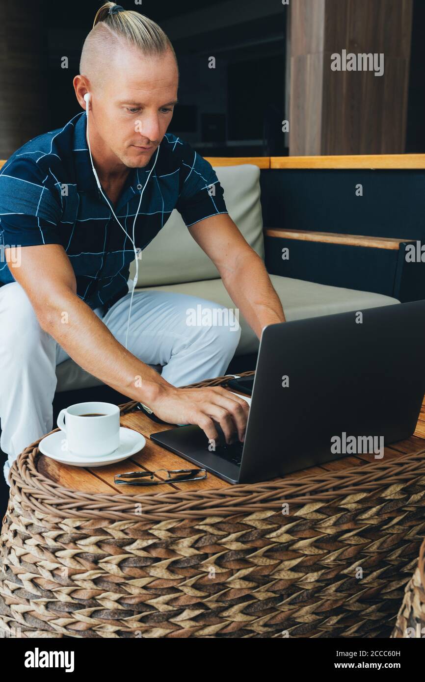 uomo d'affari in una camicia e pantaloni è in un bar, occupato con lavoro remoto utilizzando la tecnologia moderna. Funziona con un computer portatile, controlla i report o pianifica, Foto Stock