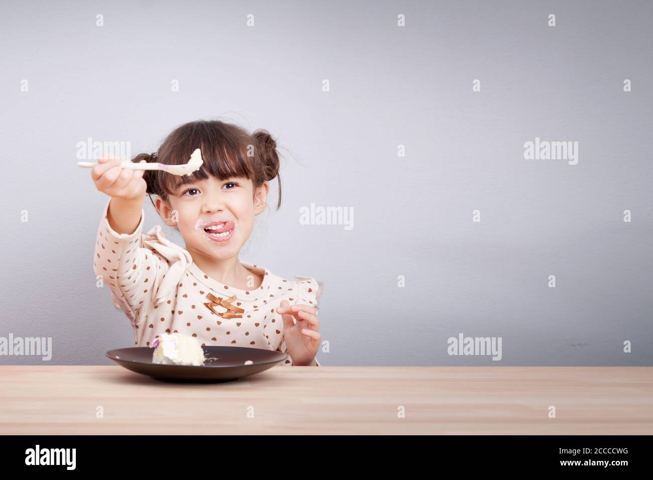 I bambini apprezzano il concetto di mangiare: Ragazza di razza mista felice piccola e carina godono di mangiare torta con faccia sorridente, bastone di lingua con cucchiaio in mano Foto Stock