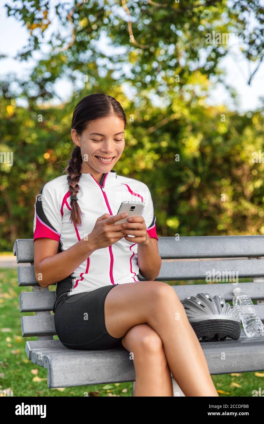 Ciclista donna che usa il telefono cellulare testando durante la pausa di allenamento al parco. Atleta asiatica ragazza in abbigliamento da ciclismo godendo di attività all'aperto in Foto Stock