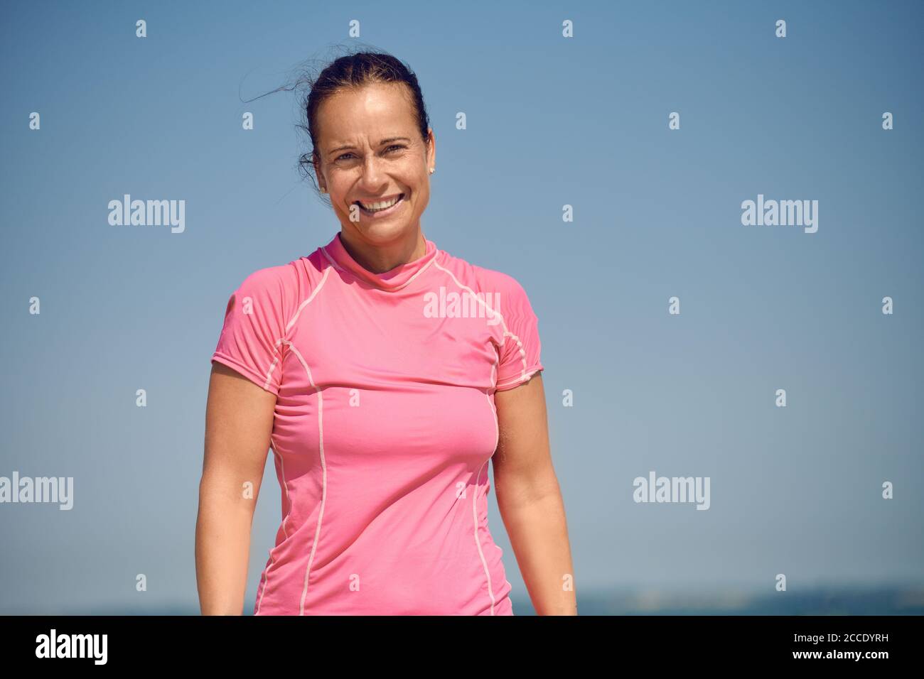 Felice sorridente attraente atletica snella donna di mezza età guardando giù la fotocamera con un sorriso caldo e amichevole contro un sole cielo estivo blu con Foto Stock