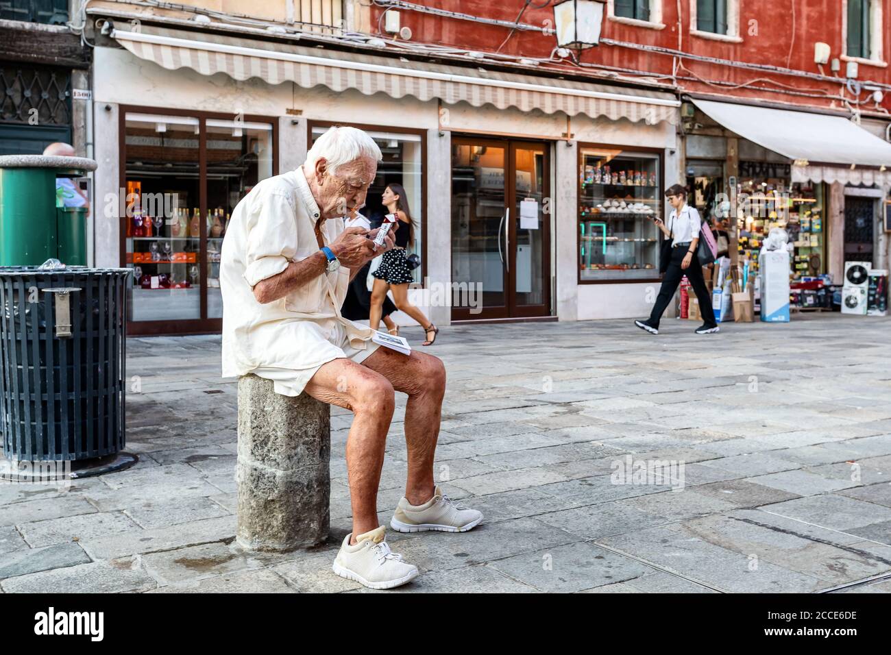Venezia, Italia - 1 agosto 2020: Un anziano seduto in strada illumina una sigaretta, mentre i turisti passeggiano per il centro della città. Foto Stock