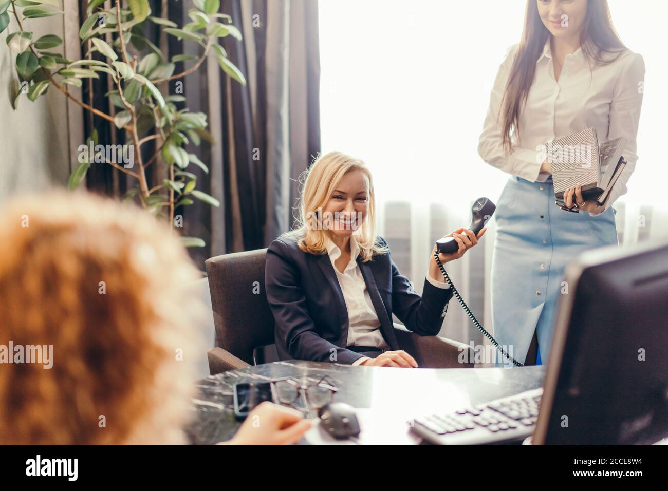 La donna bionda e amichevole Chief Executive risponde a una chiamata e ascoltando il suo collega relazione, nel suo ufficio. Business , concetto multitasking Foto Stock