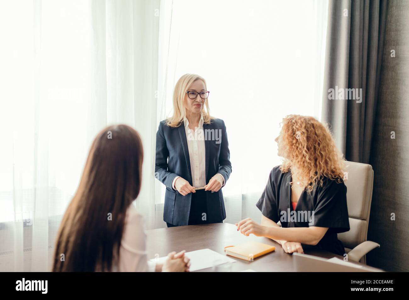 Gruppo di donne d'affari che si incontrano in ufficio. Donna bionda vestita in tuta formale che condivide le sue idee con due colleghi seduti alla scrivania. Foto Stock