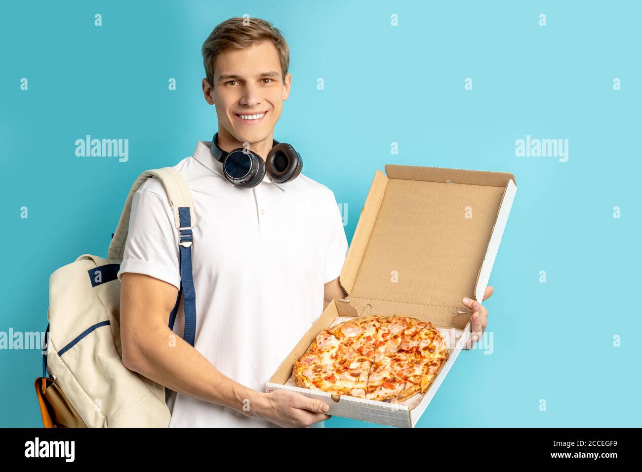 ritratto di giovane amante della pizza caucasica isolato su sfondo blu, maschio in t-shirt bianca con le cuffie tenere la pizza nelle mani, gustare mangiare, delive Foto Stock