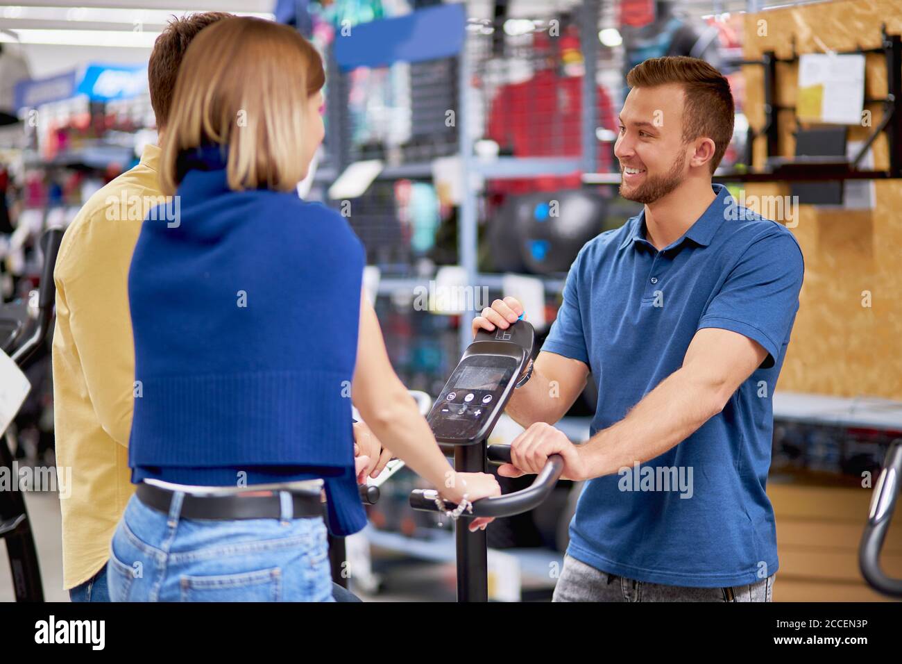 piacevole consulente aiutare i compratori in negozio di abbigliamento sportivo, consultarli e parlare di tutte le caratteristiche di attrezzatura. donna siede su cyclette, pedali Foto Stock