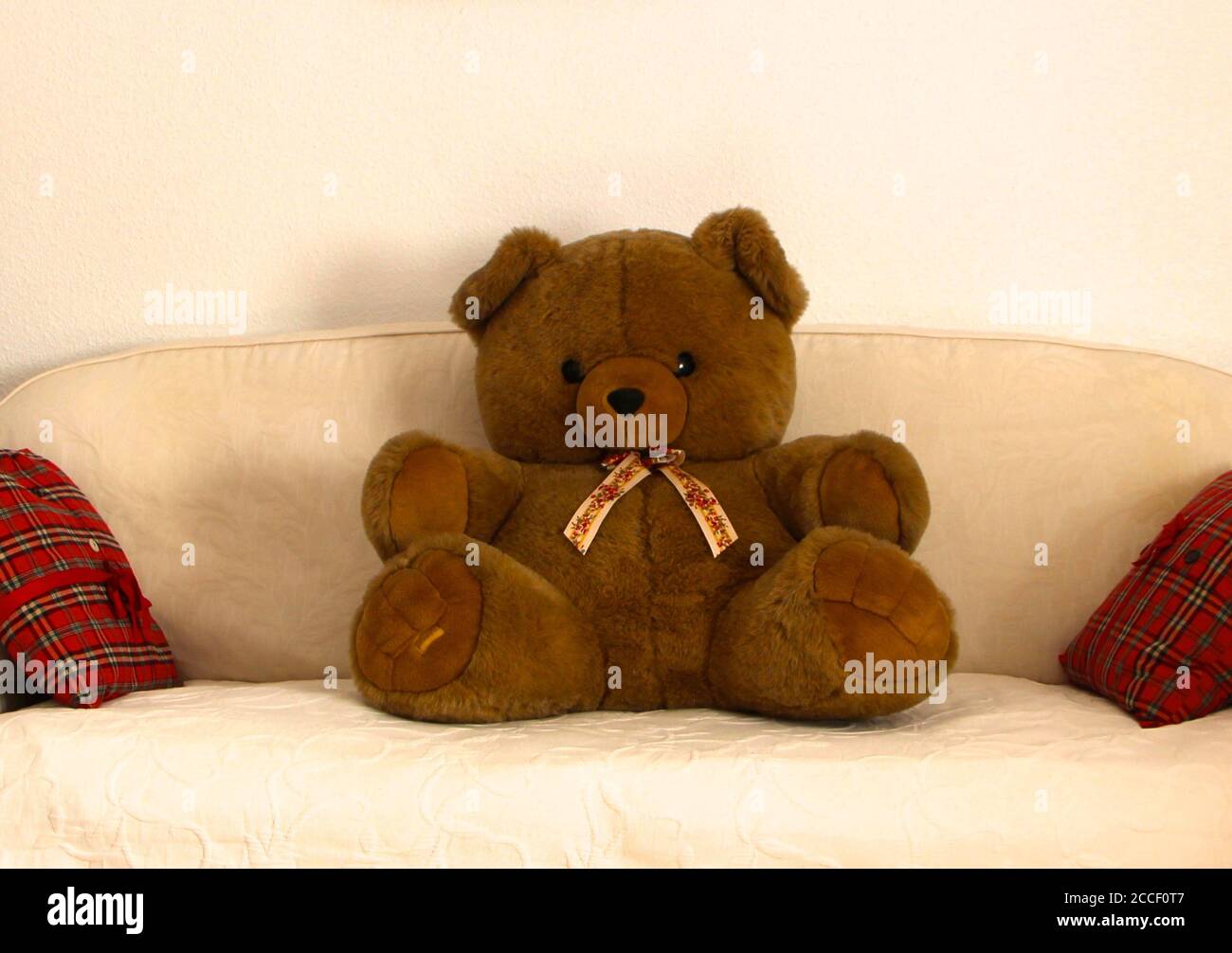 Foto di un grande orsacchiotto giocattolo marrone per bambini con un nastro di cravatta da collo seduto su un divano coperto da un tiro color crema e cuscini in plaid rosso Foto Stock
