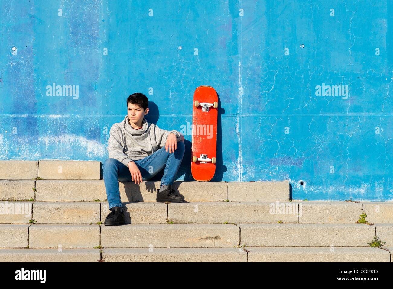 Un teen serio seduto sulla rampa di skatepark con uno skateboard, guardando via Foto Stock