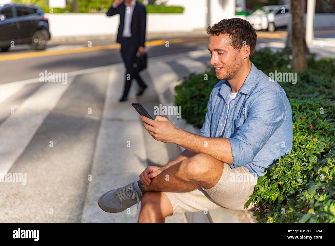 Telefono cellulare felice giovane uomo SMS SMS messaggio su smartphone in attesa di riunione sulla strada della città. Gente di stile di vita urbana all'aperto in estate Foto Stock