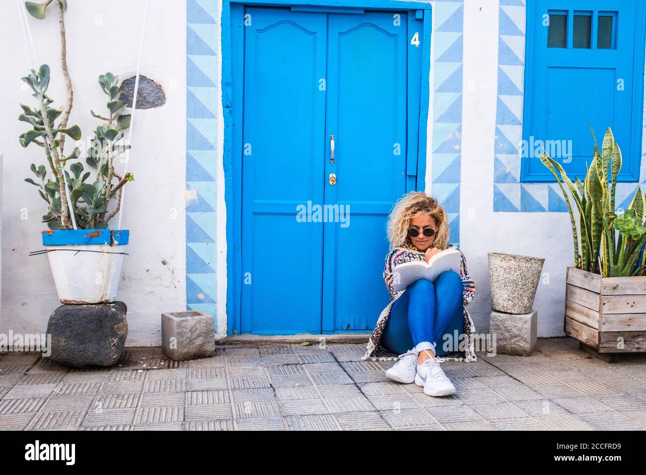 Chererful e rilassata bella donna sedersi fuori una colorata blu casa in strada leggendo un libro sul pavimento - la gente del proprietario delle proprietà domestiche Foto Stock