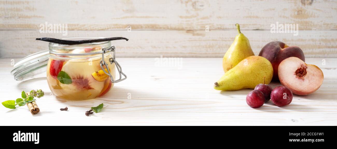 Pere, pesche e prugne di ciliegio, fresche e in scatola in un vaso di vetro con cannella, menta e vaniglia su uno sfondo di legno dipinto di bianco, panoramico Foto Stock