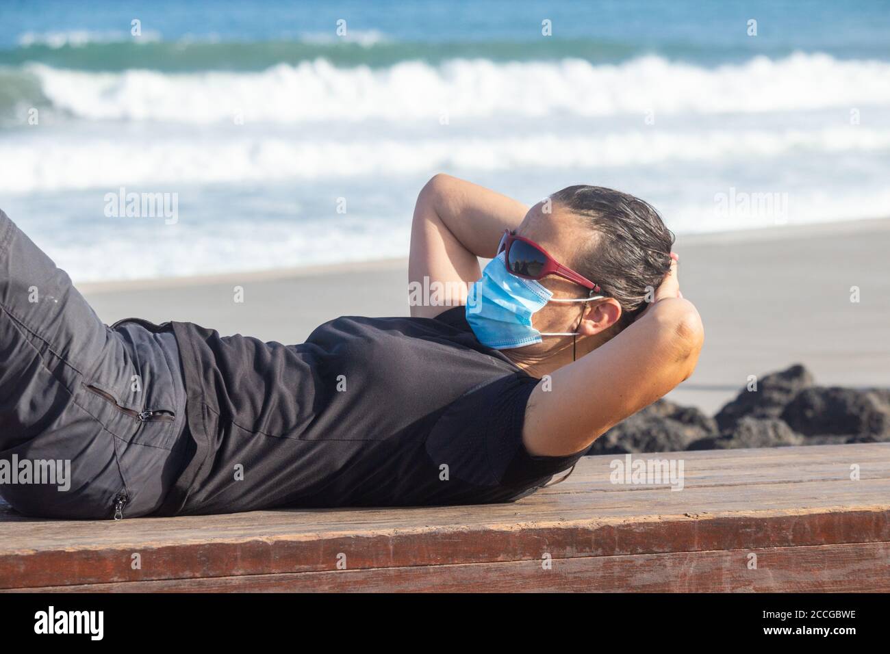 Donna che indossa il viso che si allunga sulla spiaggia durante la pandemia di Covid. Foto Stock