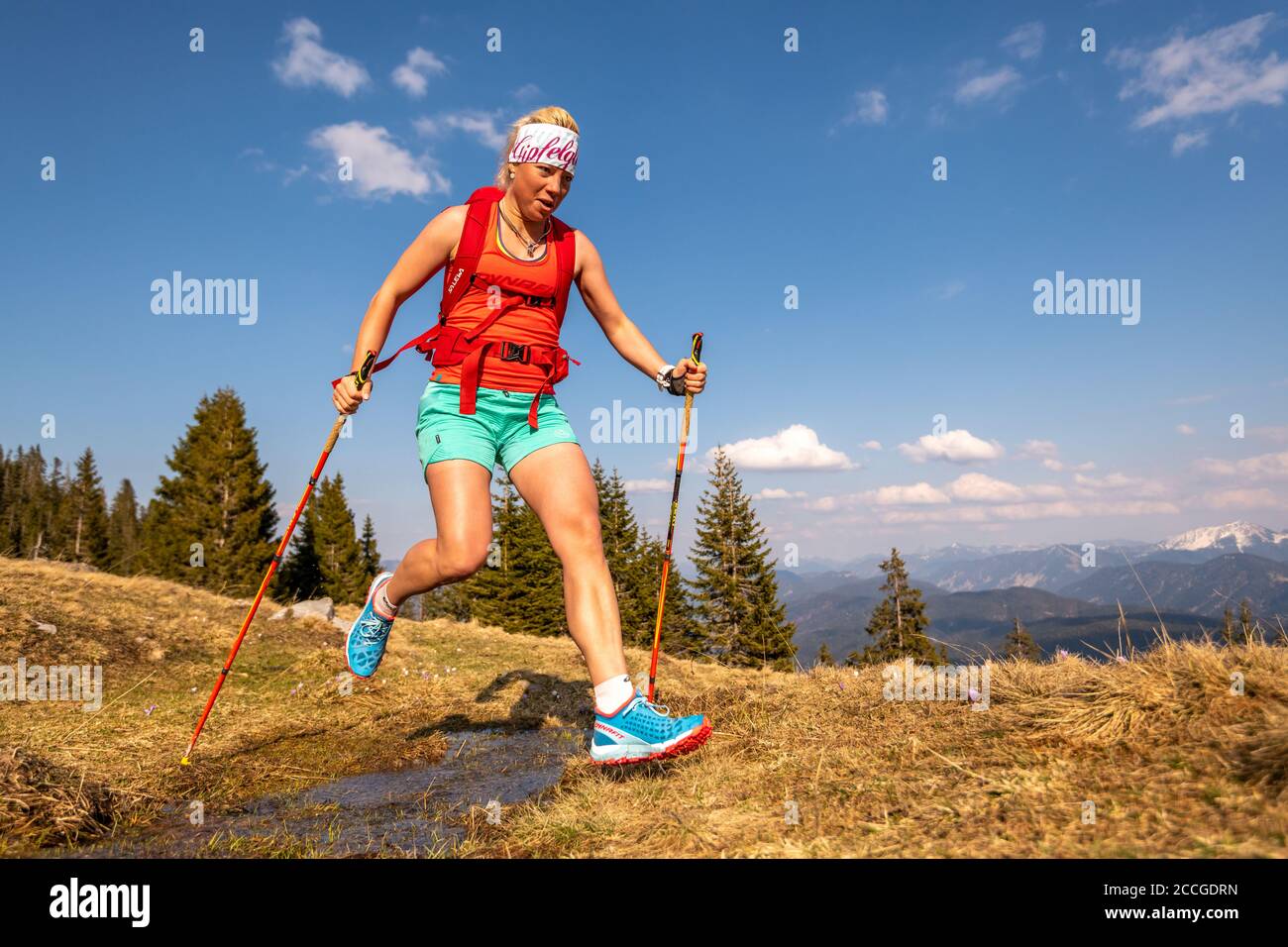 Il corridore del sentiero salta su un piccolo ruscello su un prato di montagna, sullo sfondo le Alpi tedesche e il cielo blu. Foto Stock