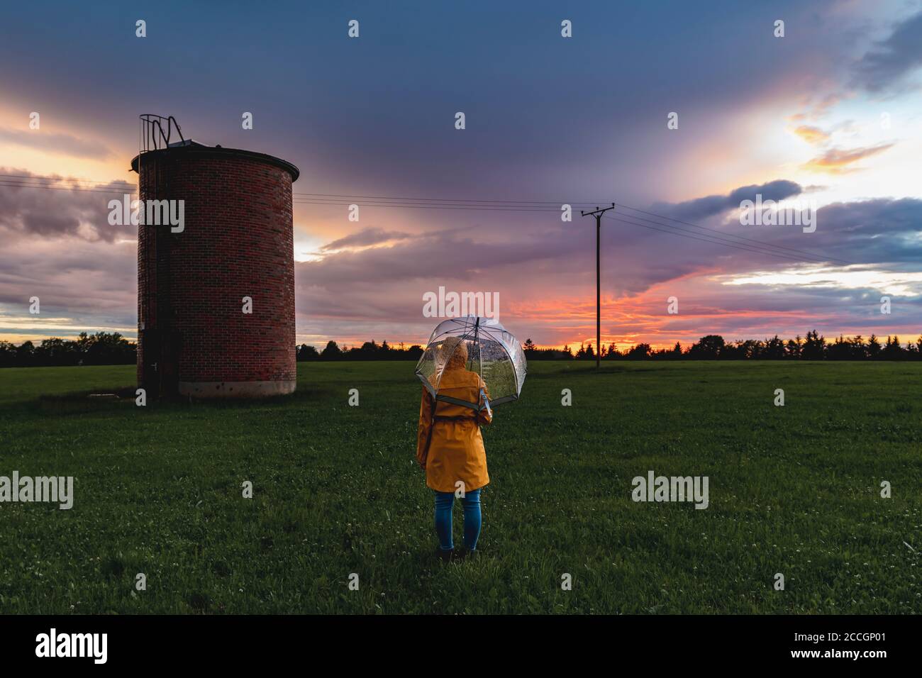 Tramonto con pioggia. In primo piano una persona con impermeabile ed ombrello e un silo di mattoni, sullo sfondo un cielo di colori spettacolari Foto Stock Tramonto con pioggia. In primo piano una persona con impermeabile ed ombrello e un silo di mattoni, sullo sfondo un cielo di colori spettacolari Foto Stock