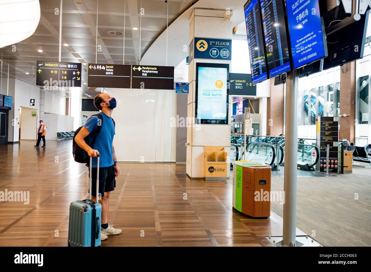 Barcellona, Spagna - 20 agosto 2020: Viaggiatore con maschera facciale in aeroporto con trolley, guardando il calendario dei voli, da solo in spazio vuoto, in viaggio Foto Stock