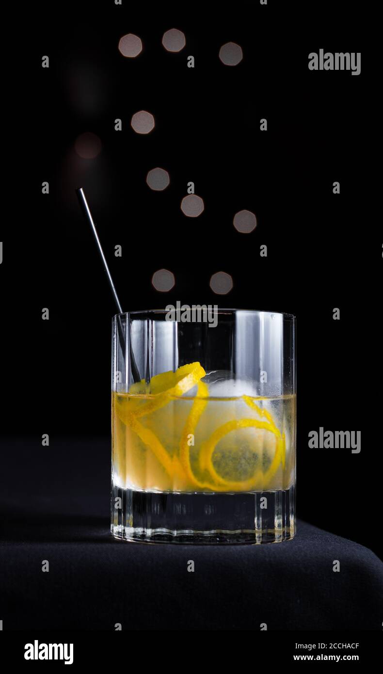 Hip, slanciato, moderno scenario di cocktail artigianali con guarnitura al limone e sfera di ghiaccio in vetro roccioso isolato su sfondo nero con cerchi in bokeh grigio. Foto Stock