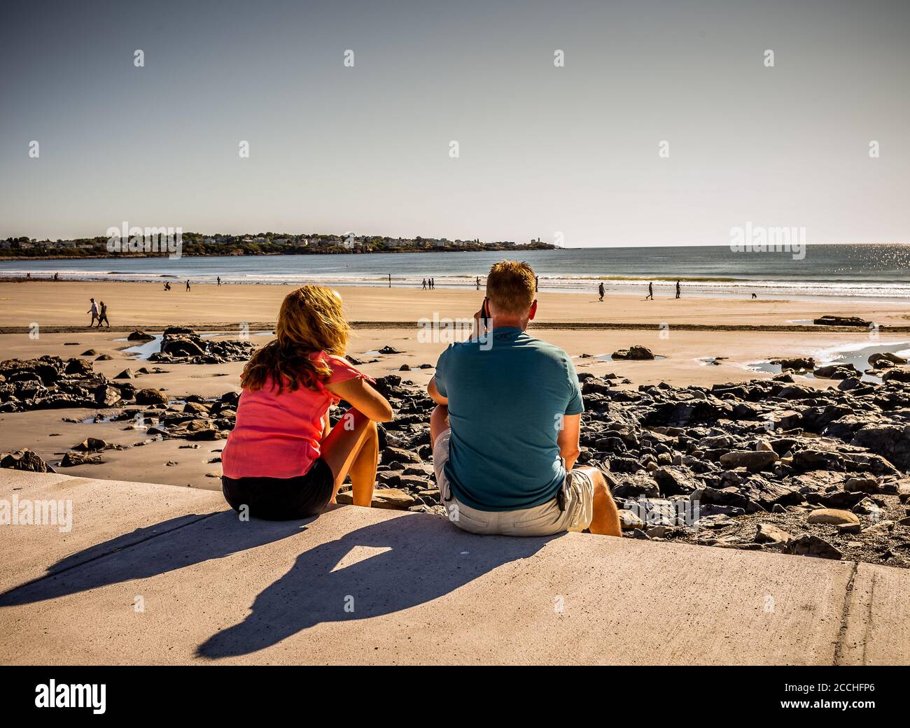 Una coppia siede del marciapiede che guarda fuori l'oceano, mentre l'uomo parla sul suo telefono, lui parla per qualche volta questo il primo giorno della loro vacanza. Foto Stock