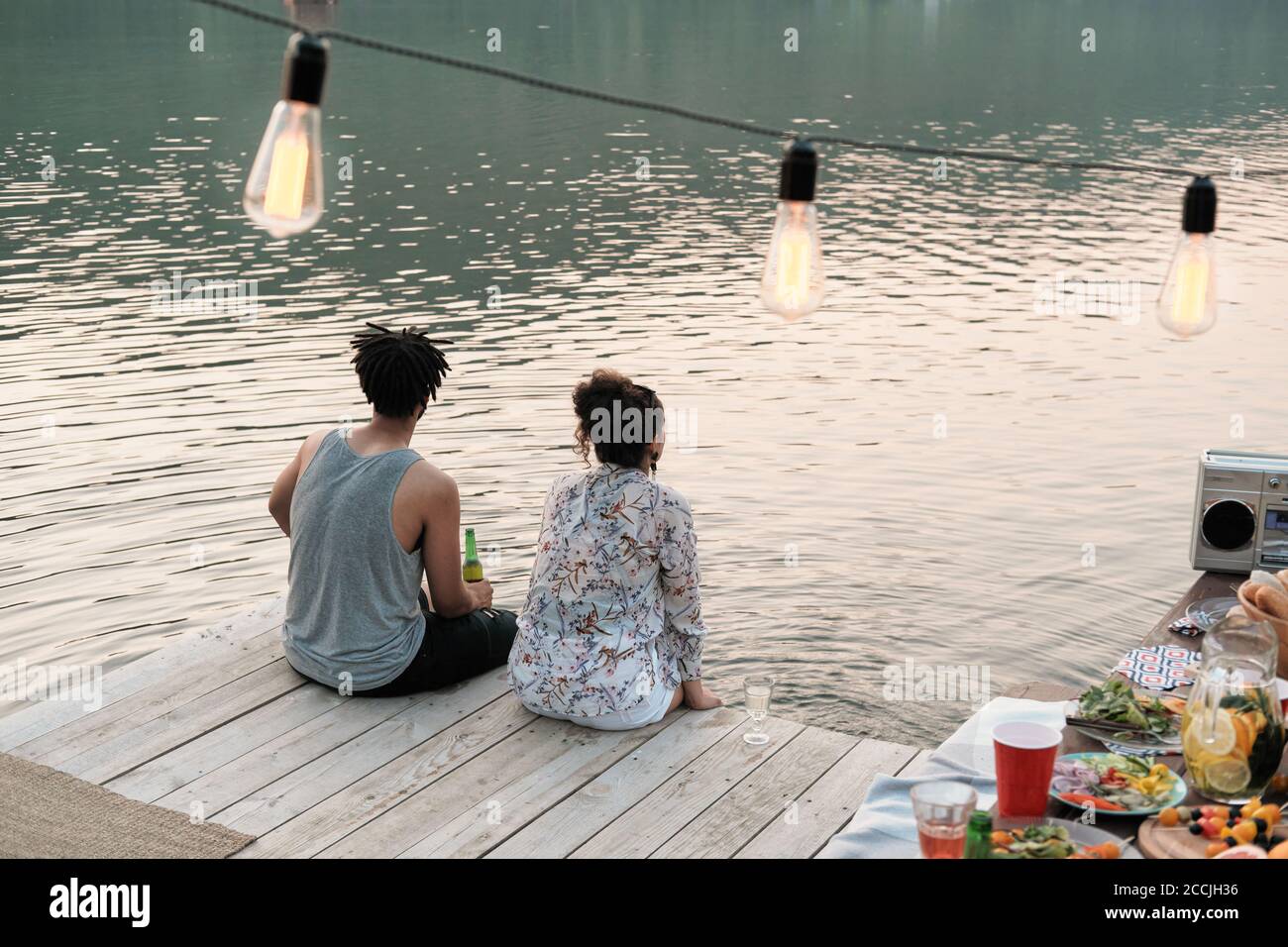 Vista posteriore della giovane coppia seduta su un molo e. guardando la vista del lago hanno una data all'aperto Foto Stock