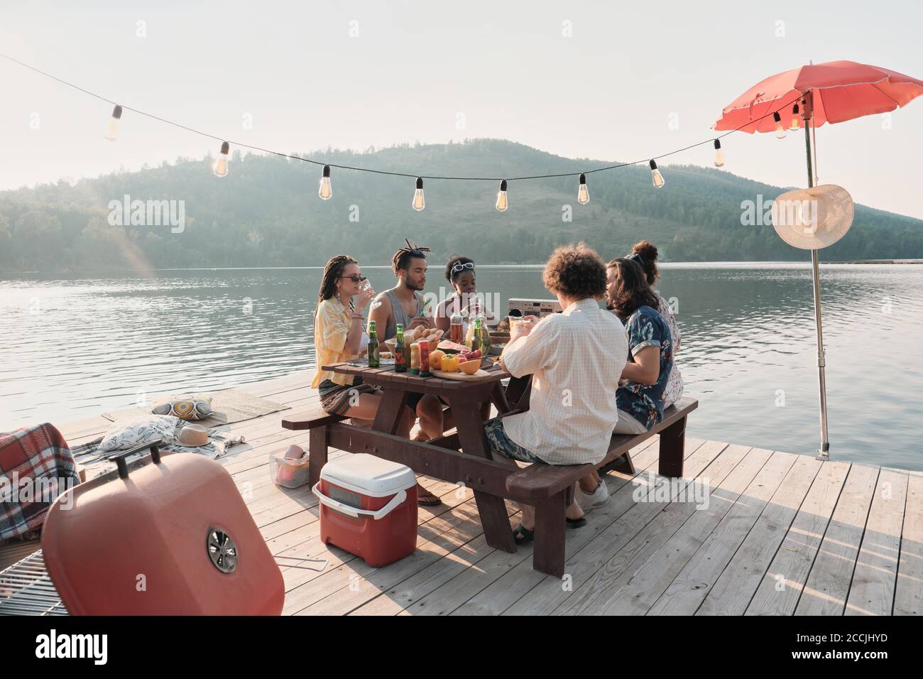 Gruppo di amici seduti al tavolo e cena su un molo con bellissimo paesaggio del lago Foto Stock