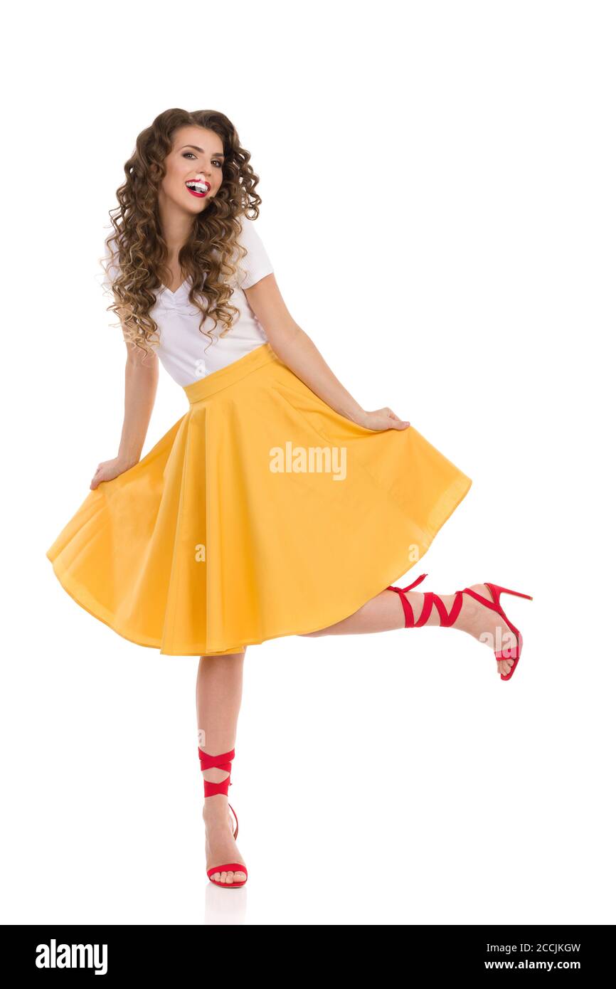 Giovane donna spensierata con gonna gialla, top bianco e tacchi alti rossi è in piedi su una gamba, ballando e ridendo. Vista frontale. Foto in studio a lunghezza intera i Foto Stock