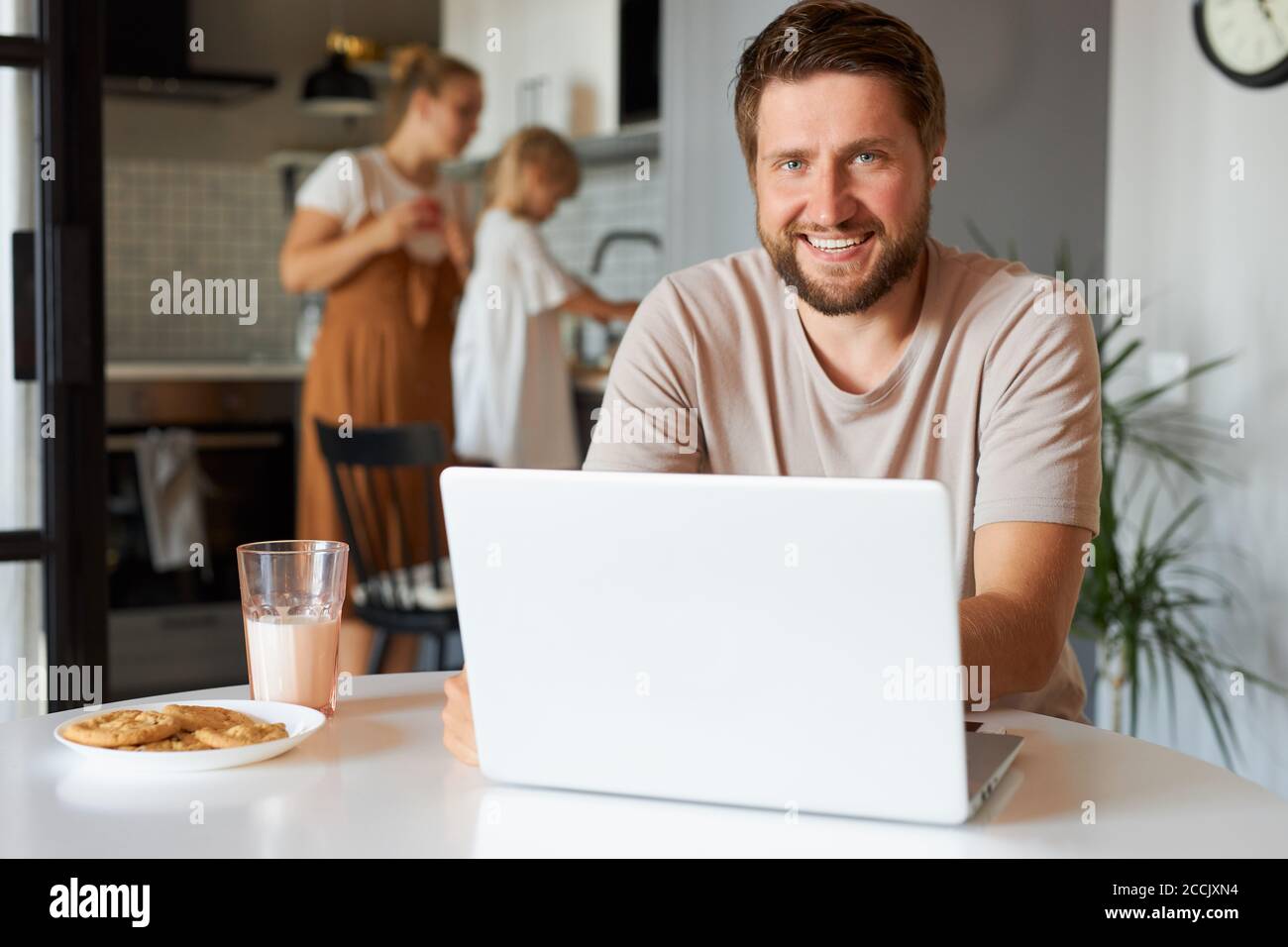 felice ragazzo positivo a freelance durante la quarantena, giovane maschio siede utilizzando laptop, donna e bambina in background Foto Stock