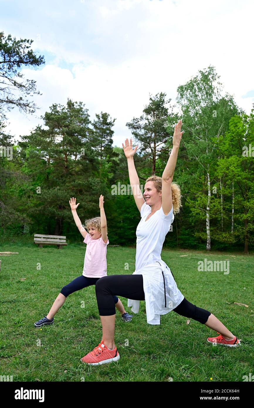 Madre con figlia che pratica guerriero 1 posa su terra erbosa in foresta Foto Stock