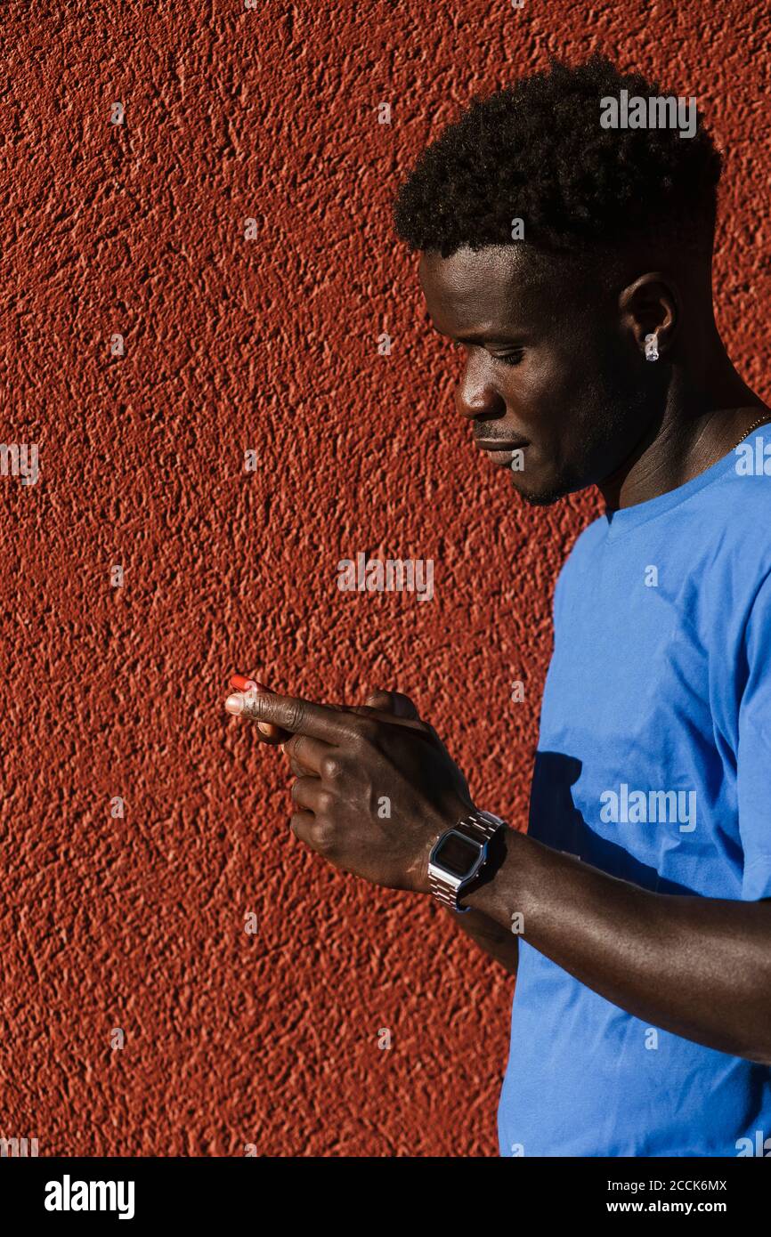 Primo piano di un giovane uomo che utilizza lo smartphone contro la parete rossa in città durante il giorno di sole Foto Stock