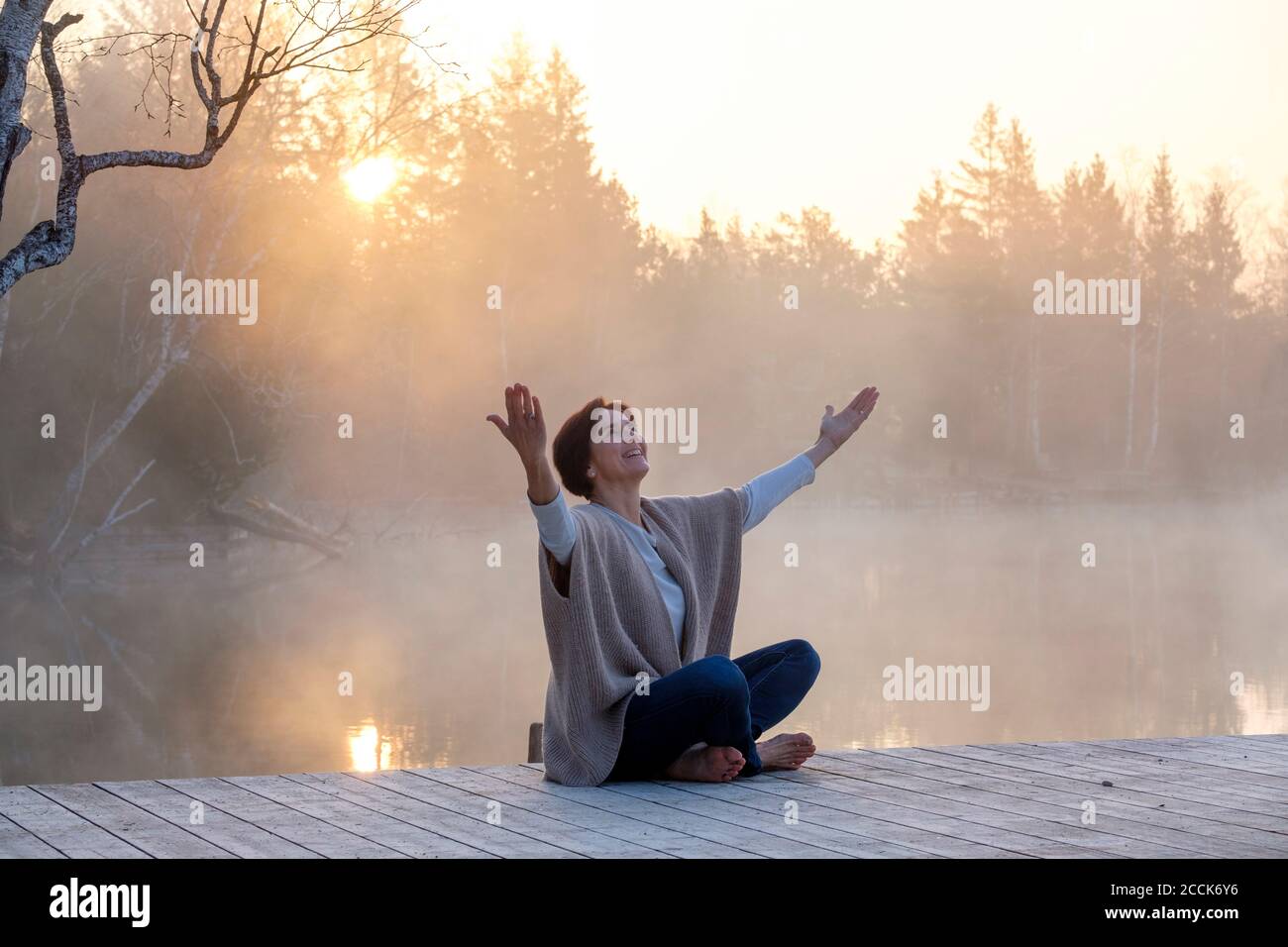 Donna adulta che medita sul molo di Lakeshore all'alba foggy Foto Stock
