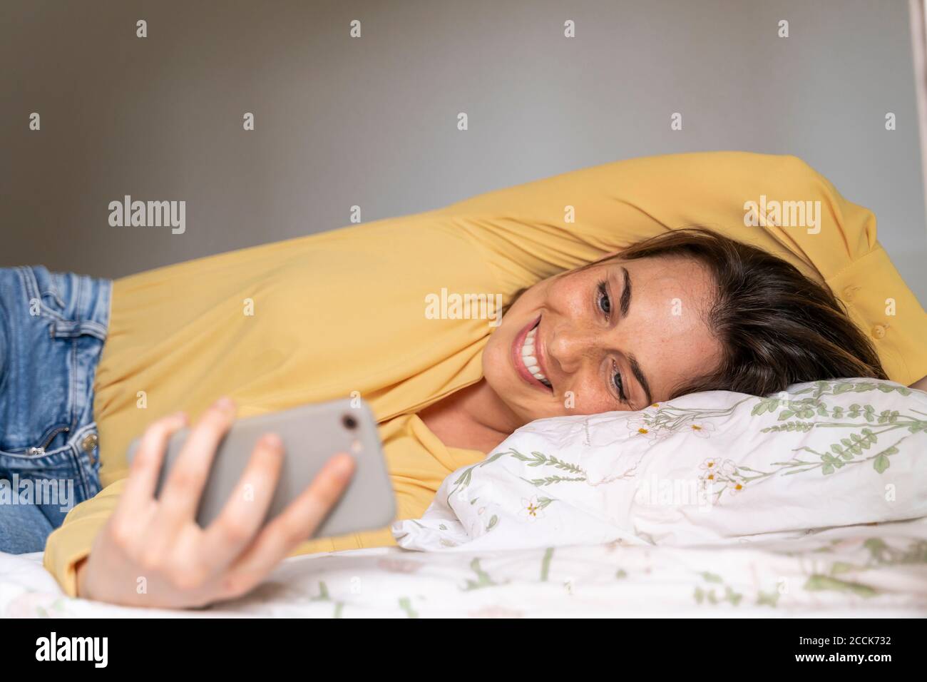 Donna sorridente che usa il telefono mentre si sdraiava in camera da letto Foto Stock