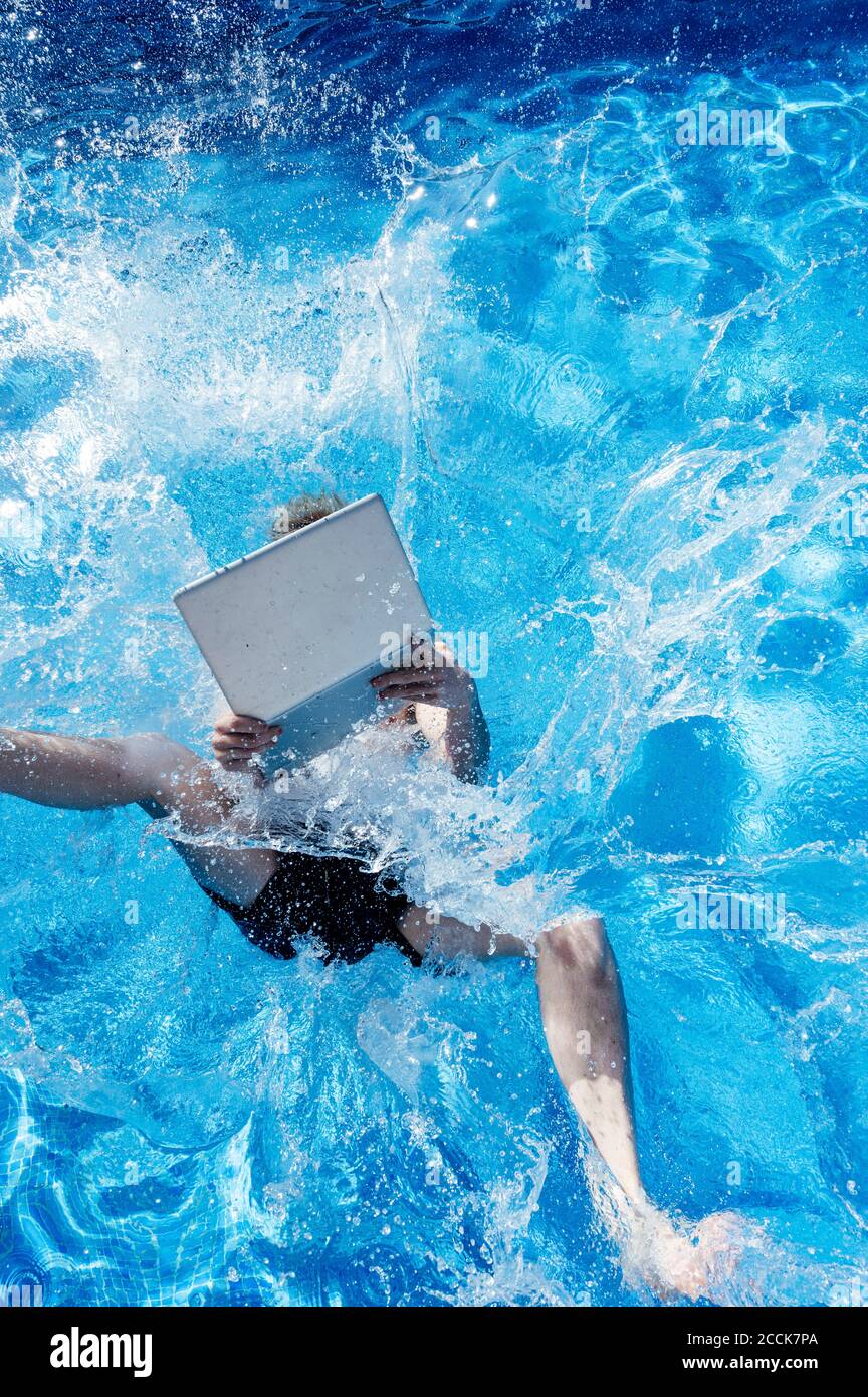 Giovane uomo con computer portatile che cade in piscina durante il sole giorno Foto Stock