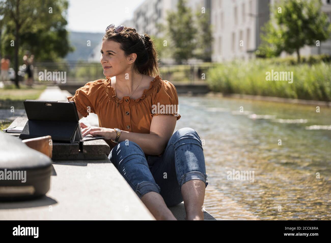Donna premurosa con computer portatile seduto accanto al laghetto nel parco durante giorno di sole Foto Stock