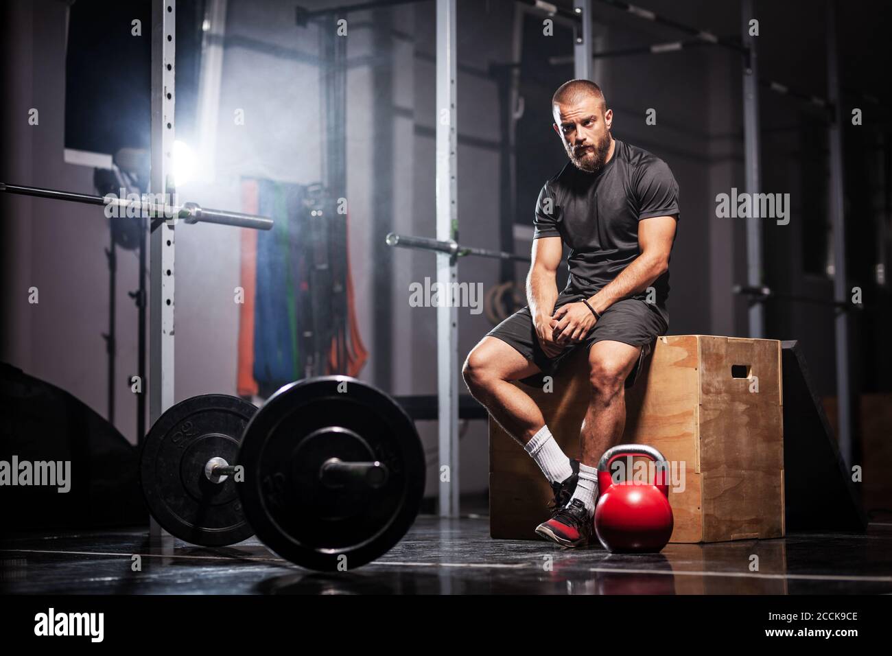 Atleta muscolare con attrezzature per il sollevamento pesi. Allenatore CrossFit in una sala fitness. Sfondo fumato. Foto Stock