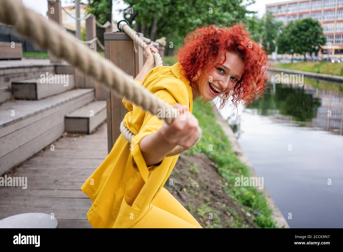 Giovane donna con capelli ricci e tuta gialla seduta sul lungofiume, che si tiene sulla corda Foto Stock