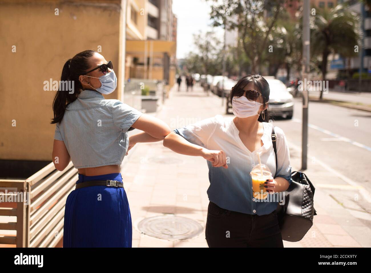 Amici femminili che indossano occhiali da sole e maschere che danno gomito scarico mentre in città Foto Stock