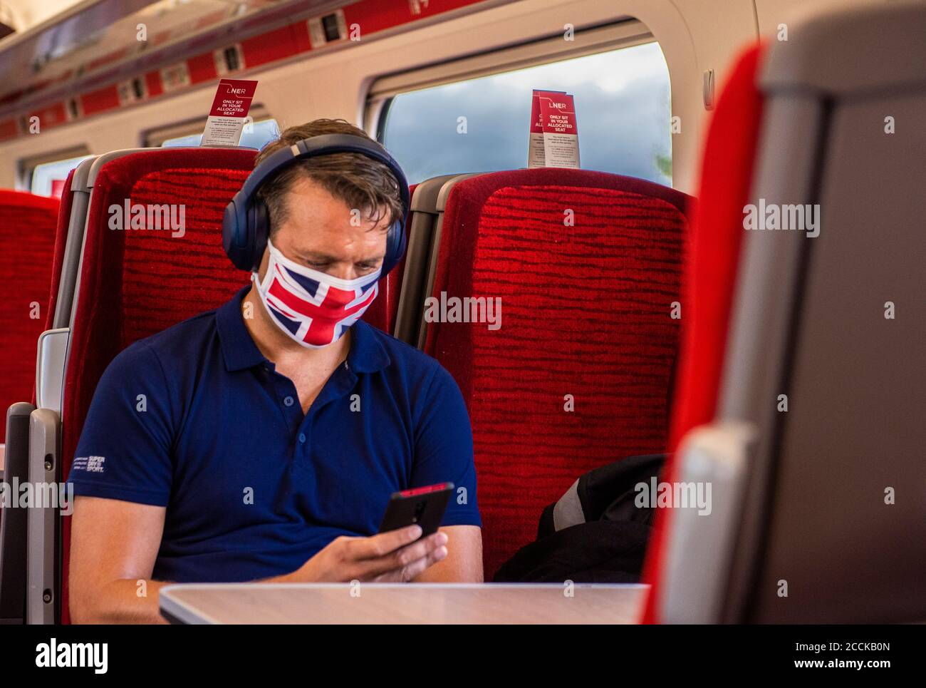Un uomo viaggia su un treno con una copertura facciale Union Jack e una polo blu sullo sfondo di sedili rossi. Foto Stock