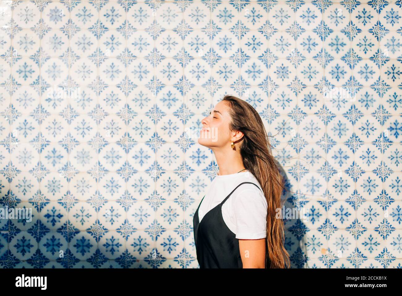 Sorridente bella donna con gli occhi chiusi in piedi da parete piastrellata il giorno di sole Foto Stock