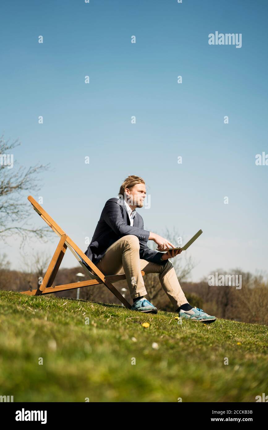 Vista a livello della superficie dell'imprenditore maschile che guarda via mentre si siede con computer portatile sulla sedia a parco durante la soleggiata giornata estiva Foto Stock