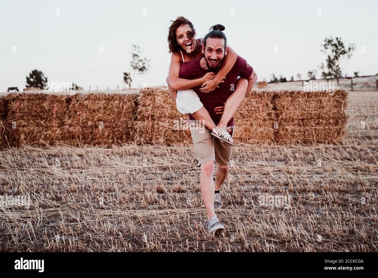 Uomo allegro che dà alla donna piggyback cavalcare contro la paglia in campo Foto Stock