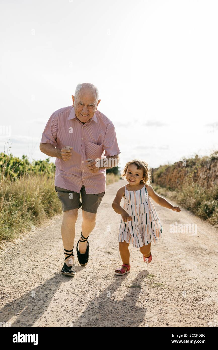 Uomo anziano giocoso che corre con la nipote sulla strada sterrata in mezzo piante durante la giornata di sole Foto Stock