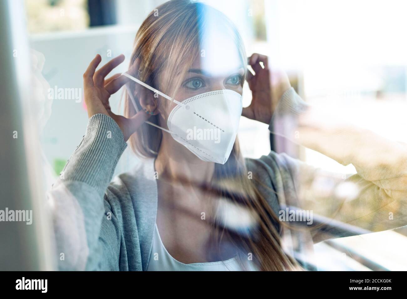 Ritratto di giovane donna che si mette su maschera protettiva dietro la finestra riquadro Foto Stock
