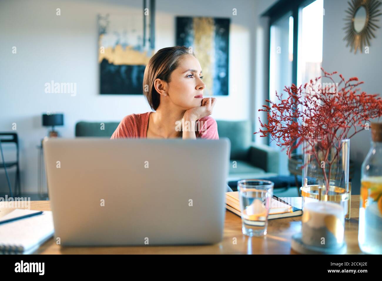 Giovane donna premurosa con un computer portatile sulla scrivania che guarda lontano seduto a casa Foto Stock