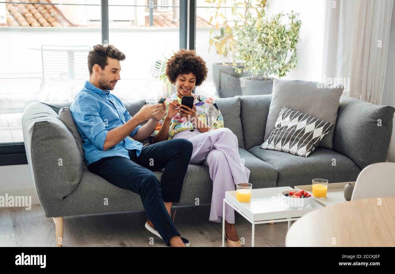 Coppia multietnica che utilizza smartphone mentre si è seduti sul divano attico Foto Stock