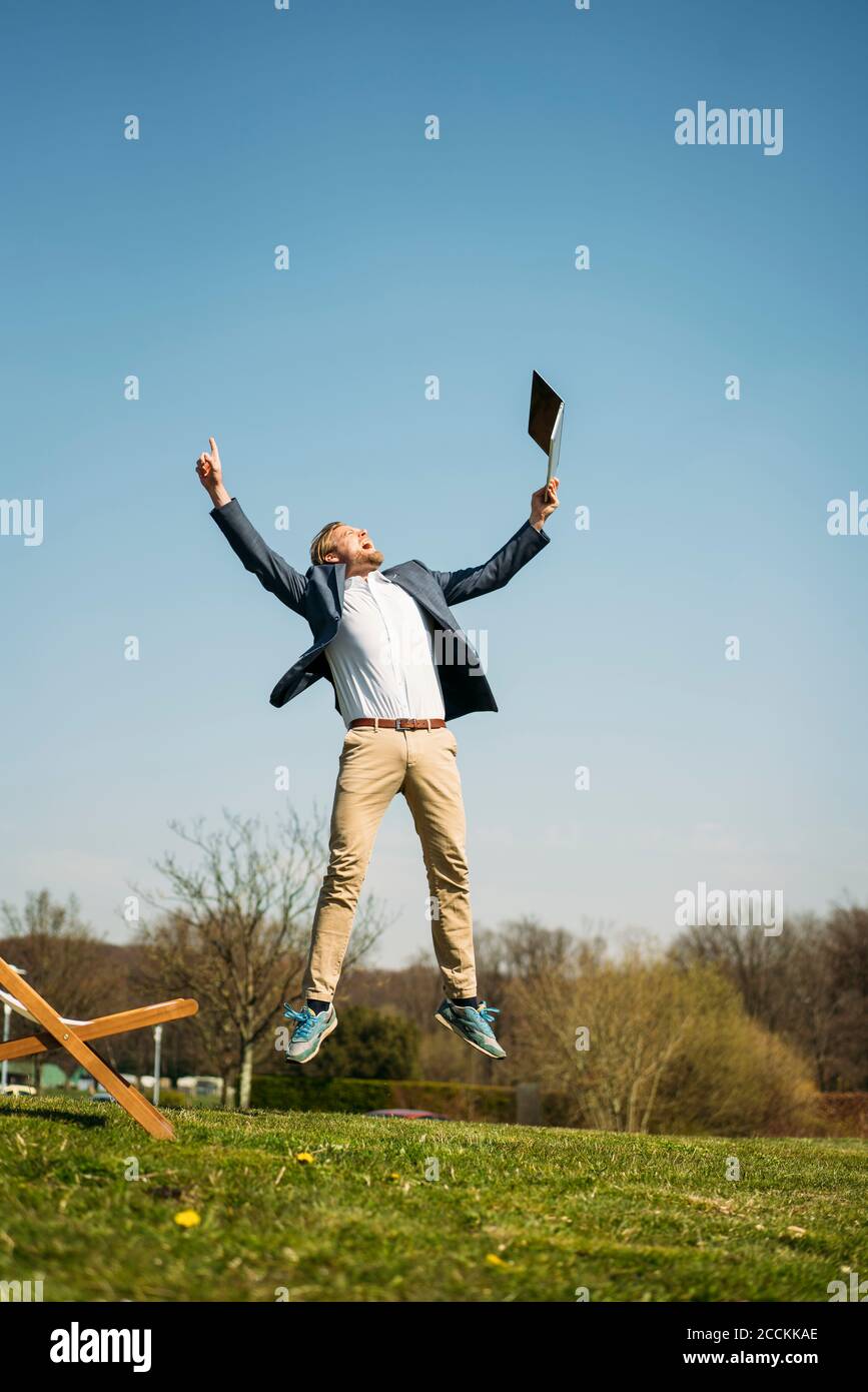 Un uomo d'affari entusiasta che tiene un computer portatile mentre salta sull'erba al parco contro il cielo limpido durante il sole giorno d'estate Foto Stock