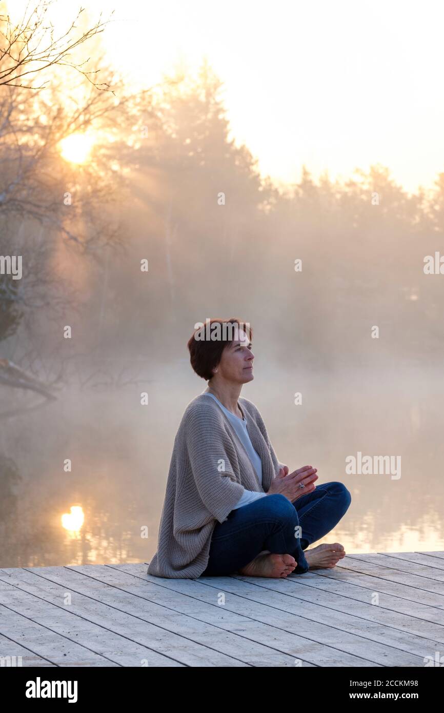 Donna adulta che medita sul molo di Lakeshore all'alba foggy Foto Stock