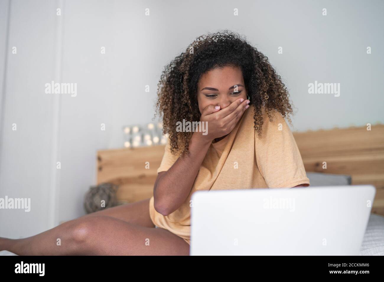 Giovane donna con la bocca che copre le mani usando il computer portatile sul letto a casa Foto Stock