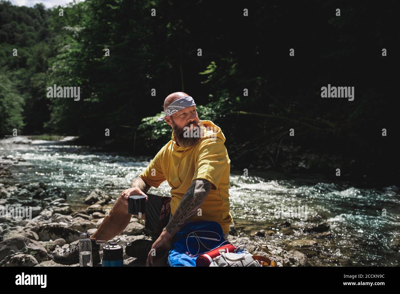 Escursionista con barba completa e felpa con cappuccio gialla durante la pausa, cucinando il tè sul lungofiume Foto Stock