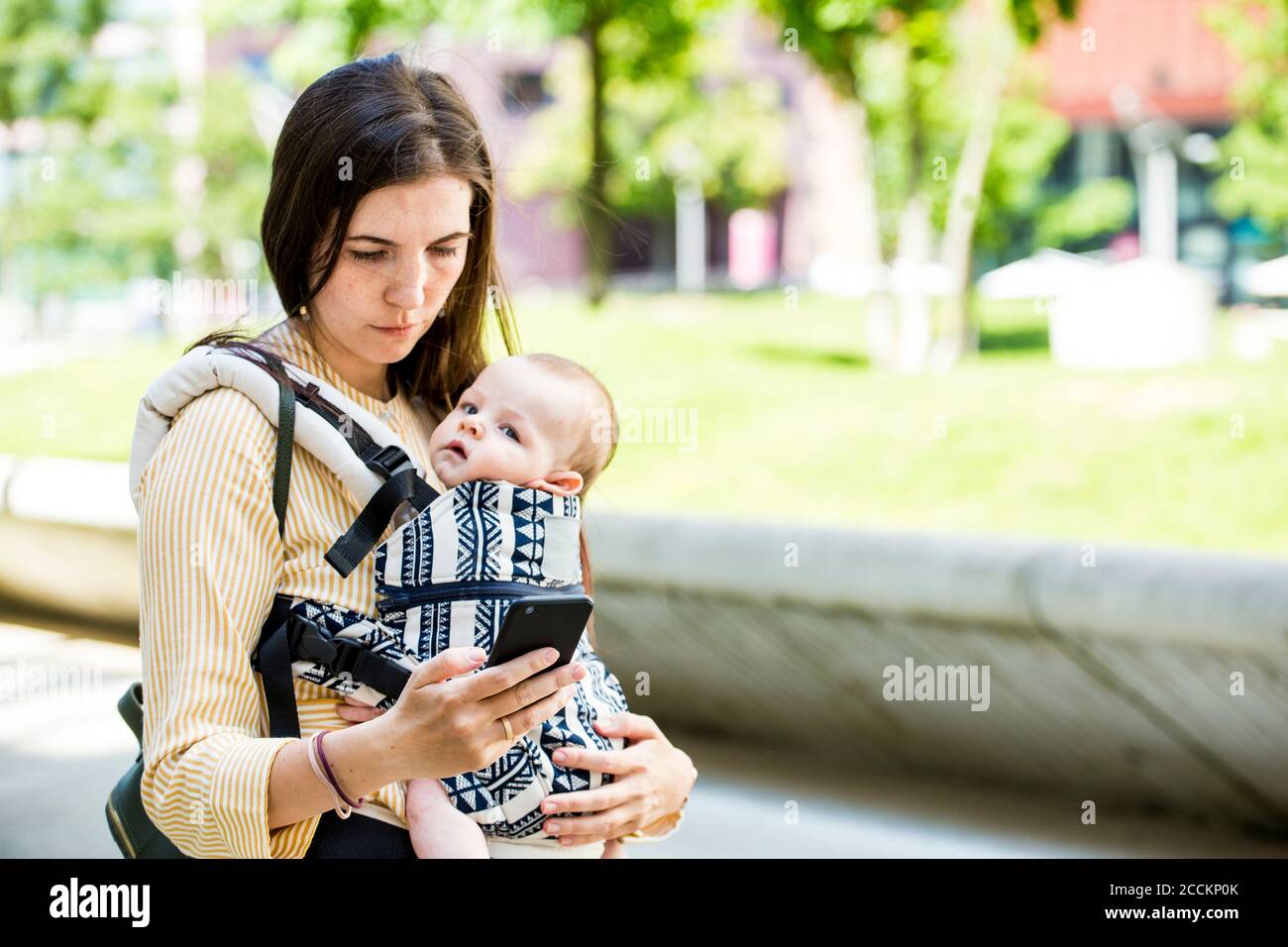 Madre con il suo bambino nel supporto del bambino usando lo smartphone in città Foto Stock