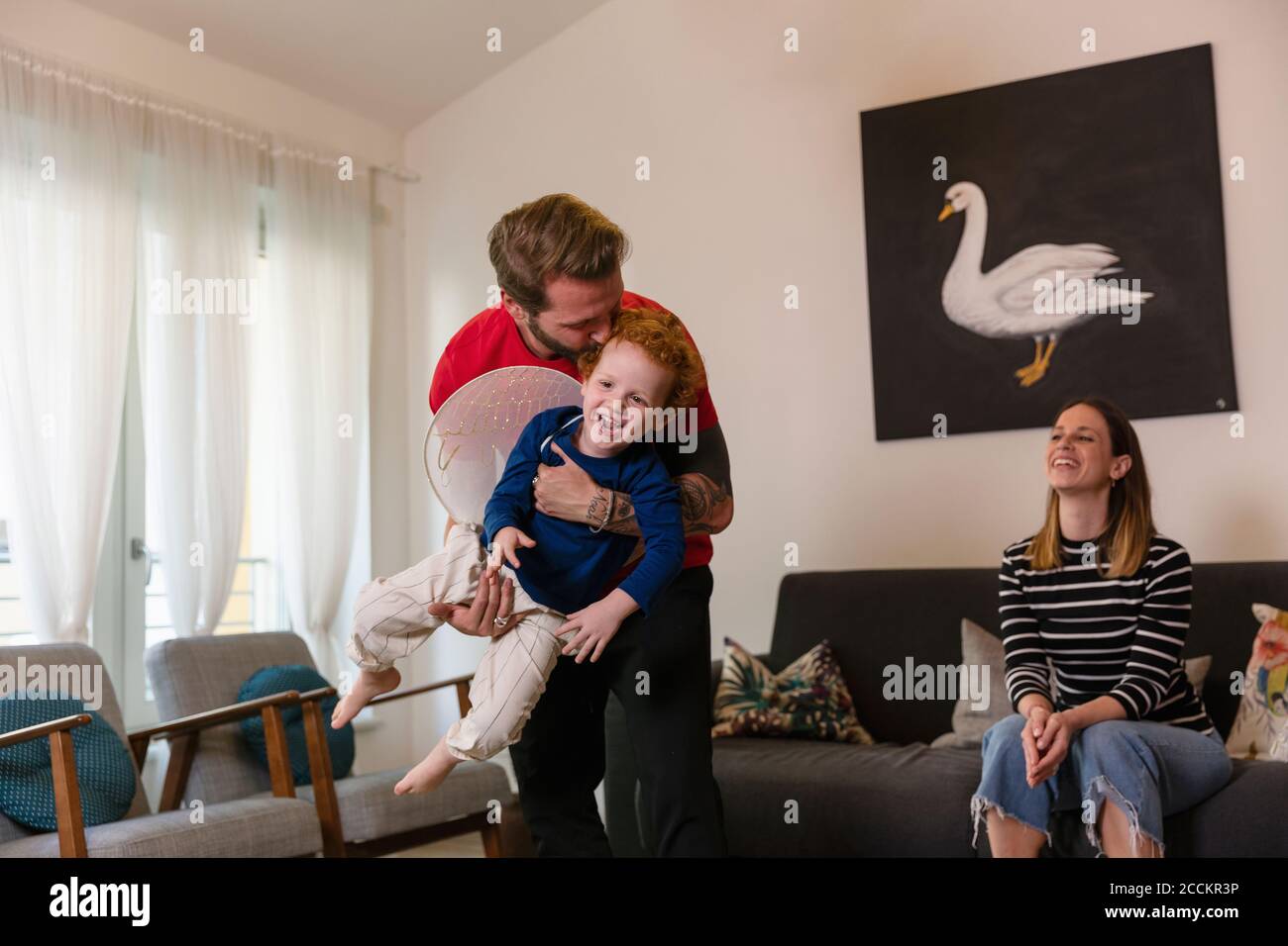 Donna felice che guarda l'uomo che porta il figlio con le ali del costume in soggiorno Foto Stock