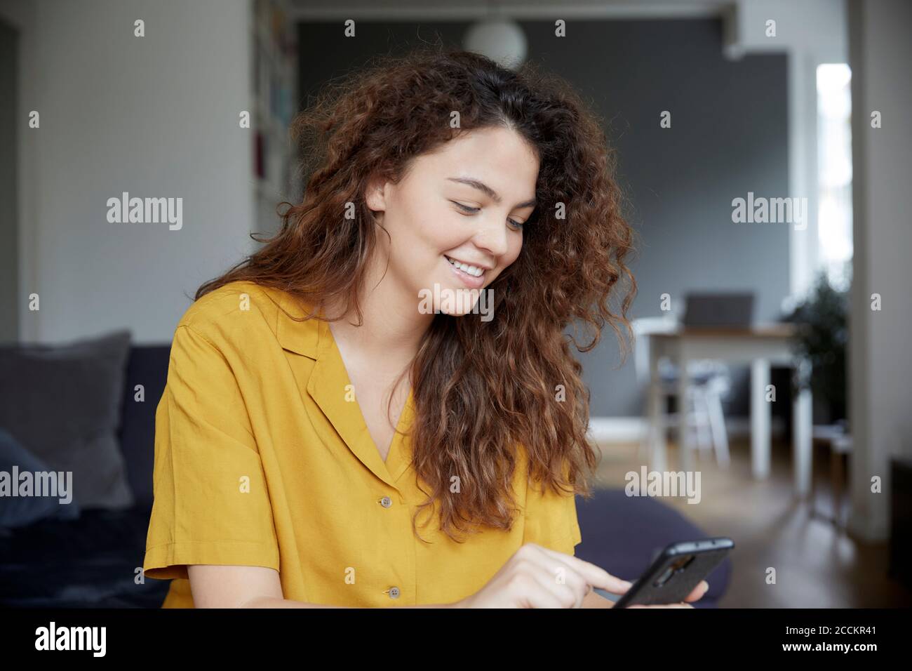 Giovane donna che sorride mentre invia messaggi di testo sullo smartphone a. casa Foto Stock