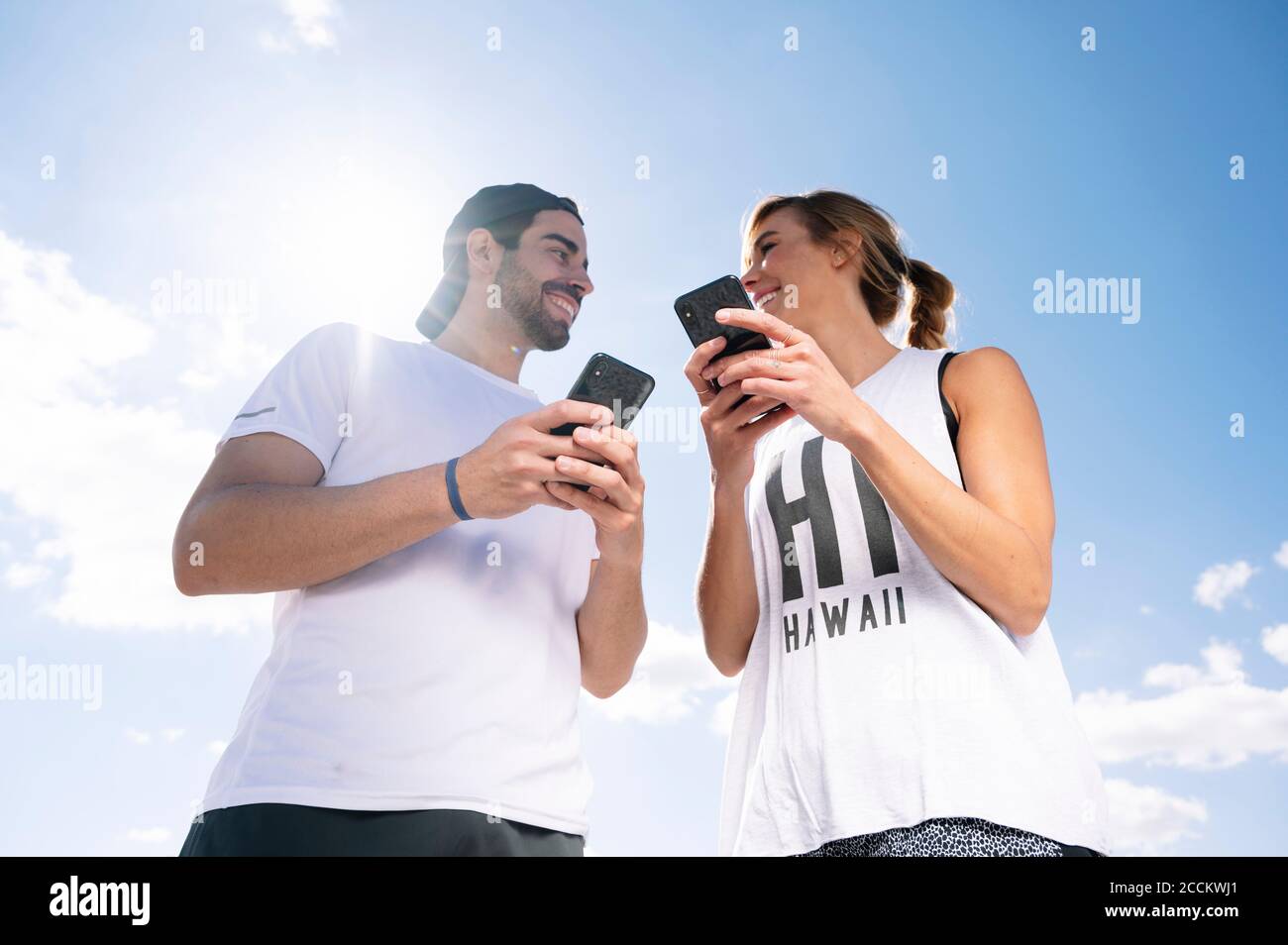 Coppia sorridente che tiene gli smartphone che si guardano l'un l'altro mentre in piedi contro il cielo durante il giorno di sole Foto Stock