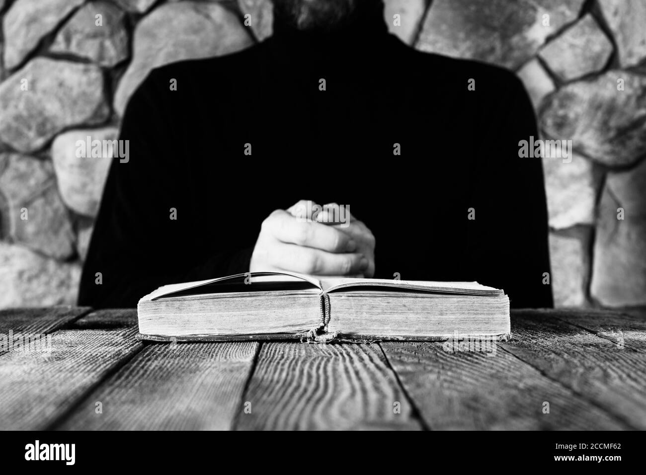 Un uomo in abiti neri davanti ad un vecchio libro aperto la Bibbia o il Corano, il concetto di preghiera, studio, stregoneria. Fuoco selettivo, bianco e nero Foto Stock